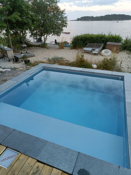 Litet rektangulärt bad med stenplattor runt om, beläget vid en strand med utsikt över vattnet och en ö i bakgrunden.