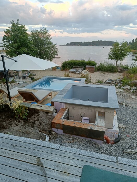 Utomhuspool med sargstenar och solstolar vid en sjöstrand, med en vacker solnedgång i bakgrunden.