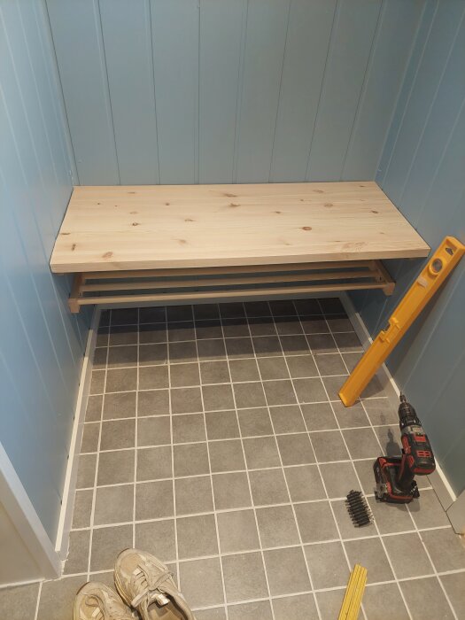 Trähylla med sittbänk monterad i ett hörn med ljusblå väggar och grått klinkergolv. En vattenpass och borrmaskin ligger på golvet.