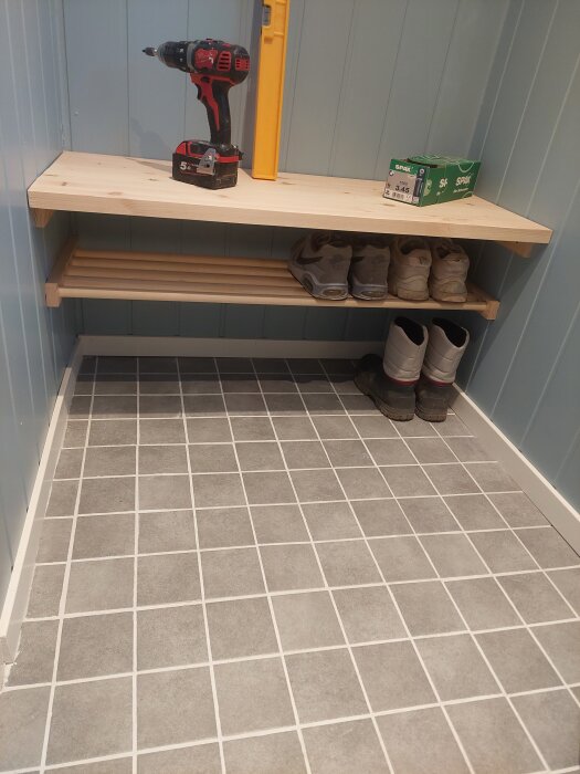 Trähylla i garage med borrmaskin, vattenpass och skor på hyllan, klinkergolv.