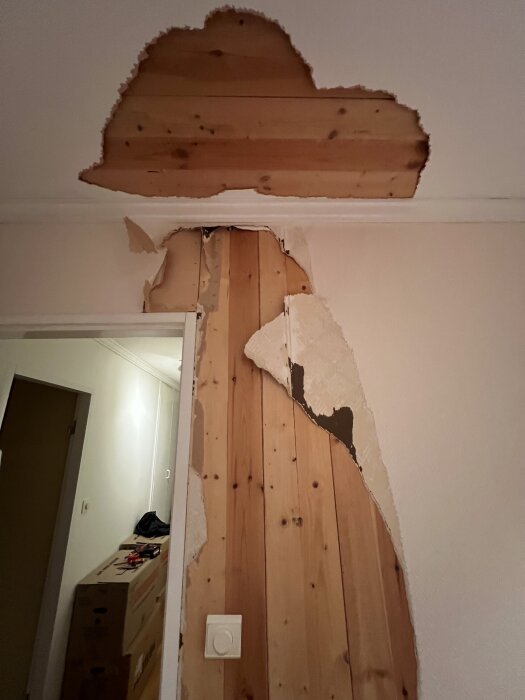 Riven väggpapp och takpapp som exponerar träpaneler, eluttag syns på väggen.