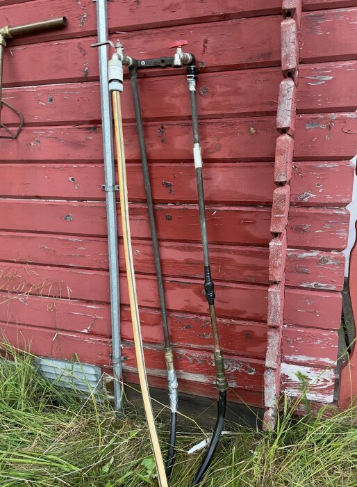 Väggmonterad vatteninstallation med metallrör och kopplingar mot en röd trävägg, med gräs vid basen.