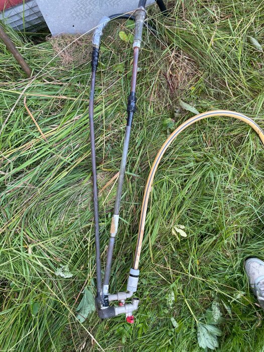 Två mjuka PEM-slangar kopplade till metallrör med trasiga kopplingar, liggande på marken med gräs och röda ventiler synliga.