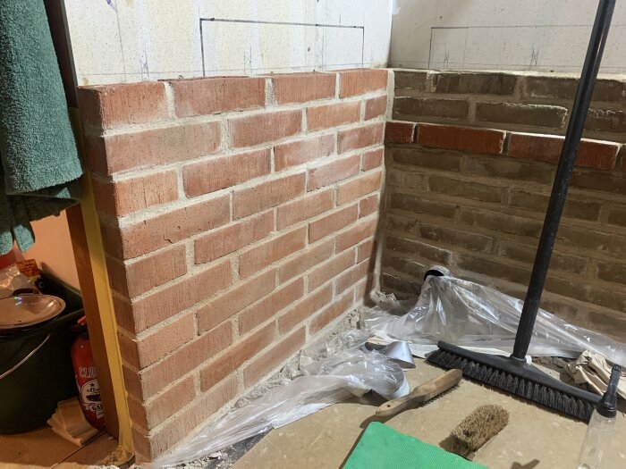 Röd tegelvägg under konstruktion med murverktyg och borstar på golvet, samt plastskydd och en handduk inomhus.