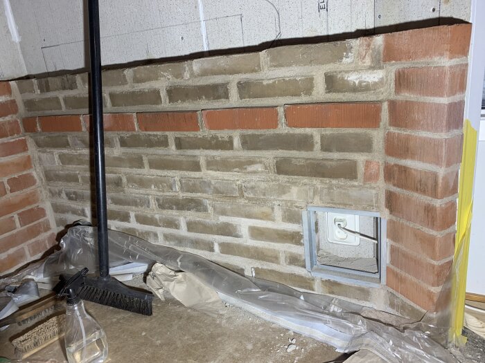 Murad brandskyddsvägg av tegelsten med synliga brukrester, sopkvast, sprayflaska och damm på golvet framför väggen. En öppen eluttag uttag syns.