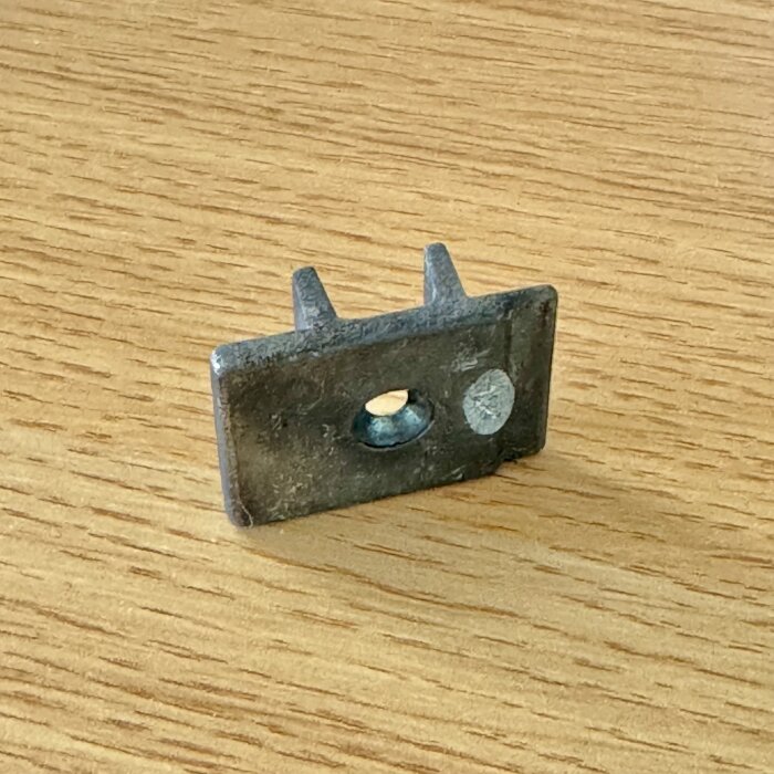 Metallfäste för fönsterspärr, rektangulär med två tubulära förlängningar och ett hål i mitten, placerad på träyta.