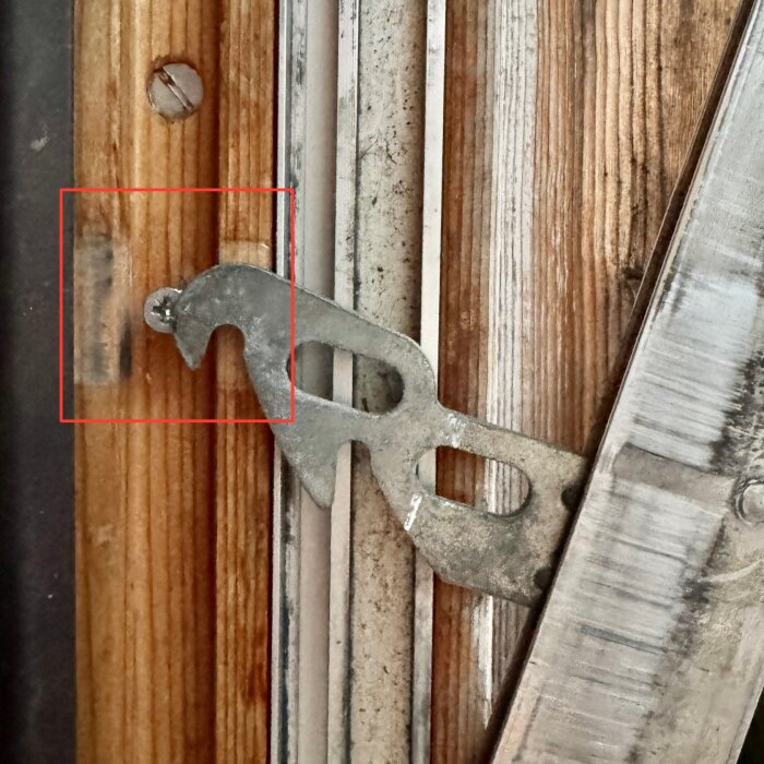 Låsdetalj för vädringsspärr på ett träfönster med metallkomponenter, närbild visar slitna delar och skruvhål.