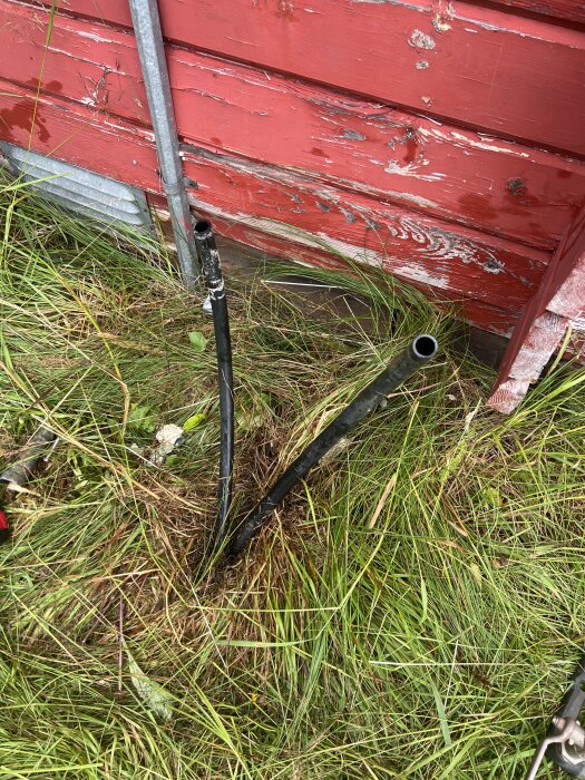 Rör och stänger i marken framför en röd trävägg, omgivna av långt gräs.