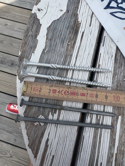 Två skruvar och två spikar liggande bredvid en tumstock på ett slitet träbord, för jämförelse inom byggprojekt.