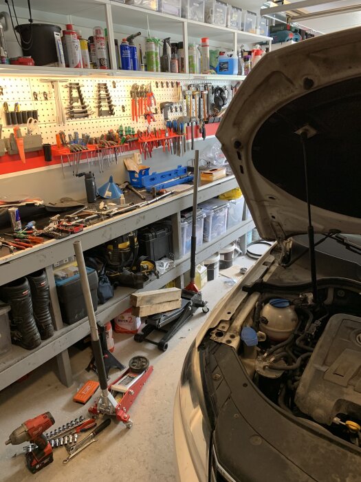 Bilens motorhuv är öppen i ett garage fullt av verktyg och utrustning, med fokus på bilreparation.