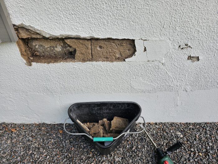 Skadad puts på garagets vägg visar sprickor och blottad tegelsten. En hink med bruten puts står på marken, omgivet av grus och en borrmaskin.