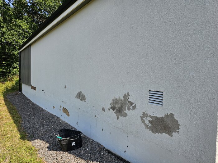 Garagets vägg med avflagad färg, sprucken puts och ventilationsgaller. Svarta hinkar i förgrunden och grus vid sidan av väggen.