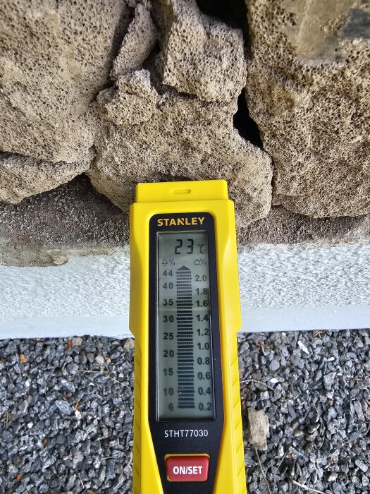 Fuktmätare visar 23.7% mot sprucken puts på garagevägg, synlig sten bakom, grus nedtill.