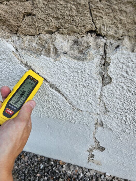 Hand håller en fuktmätare nära sprucken puts på garagevägg, visar ett område med exponerad sten och mögel i söndrigt murverk.
