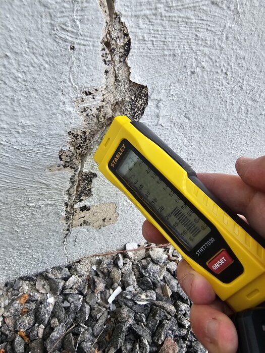 Gul fuktmätare används mot sprucken putsad vägg ovanför grus.