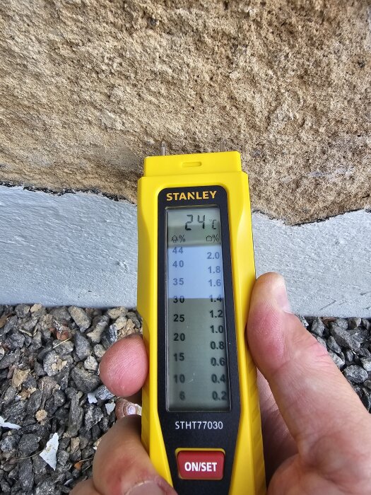 Gul fuktmätare mot murad vägg visar 24,1°C. Händer håller verktyget bredvid skrovlig puts och grus på marken.
