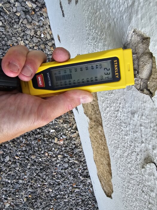 Hand håller en gul fuktmätare mot sprucken puts på garagevägg.