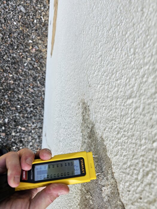 Fuktmätare med gul hölje mäter fuktighet på putsad garagvägg med flagnad färg och sprickbildning, visar värde 23% på displayen.