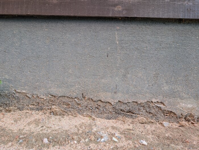 Garagesockel med avflagad puts och synlig isolering, intill sandig mark.