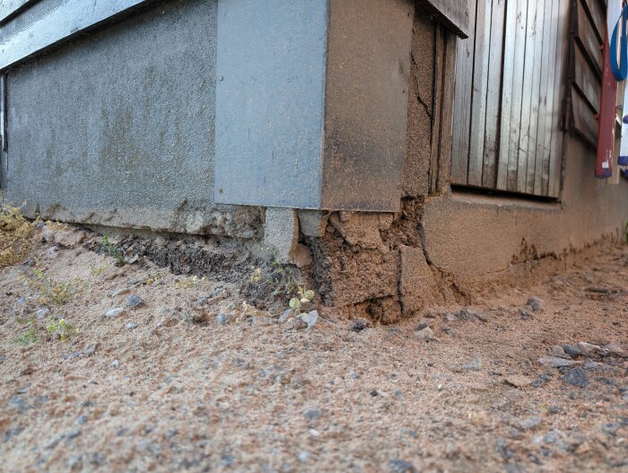 Garagesockel med sprucken puts och synlig isolering på en sandig grund bredvid ett trägarage från 60-talet.