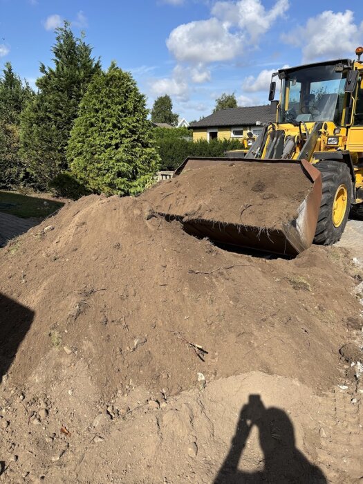 Grävskopa fylld med jord bredvid en stor jordhög, omgiven av buskar och träd i en trädgård.