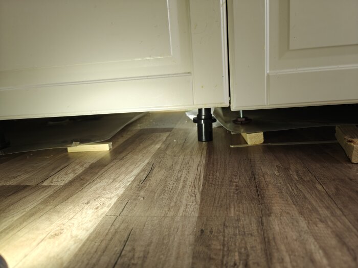 Golvplanka har rest sig vid skarv, synligt med plastskydd och träklossar under diskmaskin i kök.