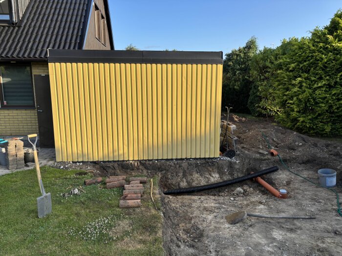 Gul byggnad bredvid gräsmatta; grävarbete med dräneringsrör synliga, en spade och tegelstenar ligger på marken.