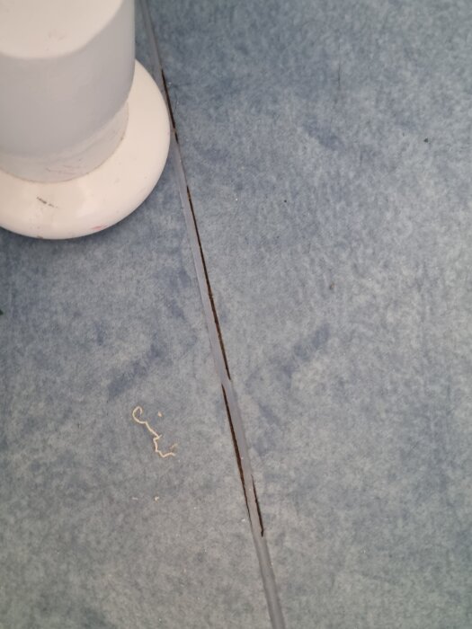 Trasig blå badrumsmatta intill ett vitt rör, med en spricka längs kanten.