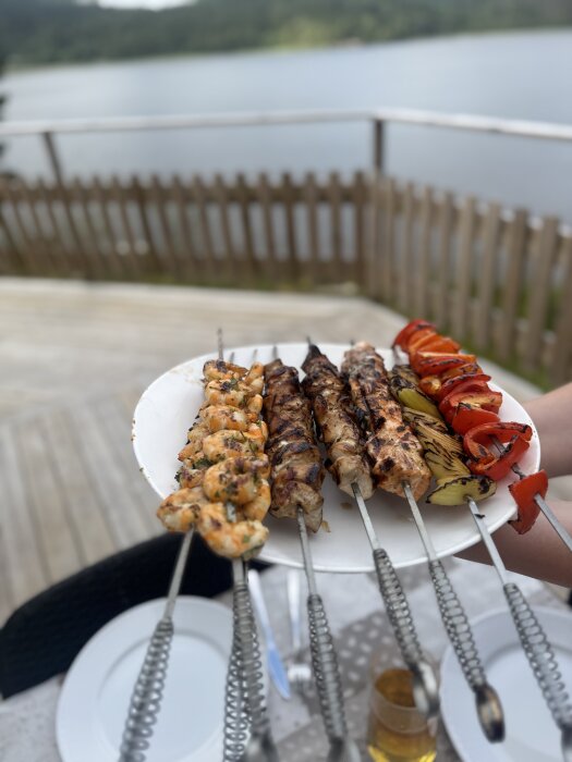 Grillspett med olika kött och grönsaker samt en bakgrund med sjöutsikt.
