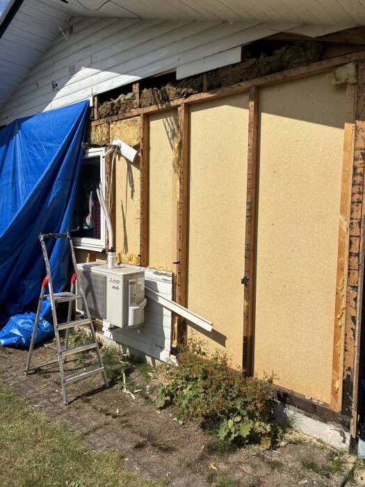 Riven gavelvägg med exponerade reglar och isolering, blå presenning täcker delvis, stege och luftvärmepump synliga intill husfasaden.
