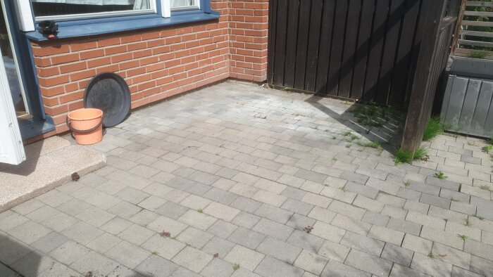 Stenbelagd yta vid radhusvägg med synliga mindre sättningar och växtlighet. En orange hink och ett svart lock står vid väggen.