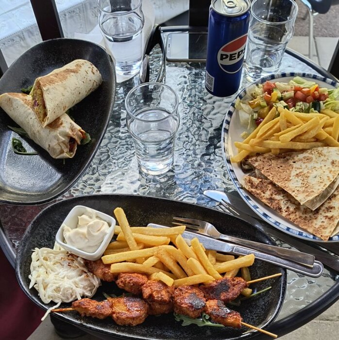 Matbord med kebabspett, pommes, wrap, coleslaw, quesadillas, sallad och Pepsi på ett glasbord i Bollnäs.