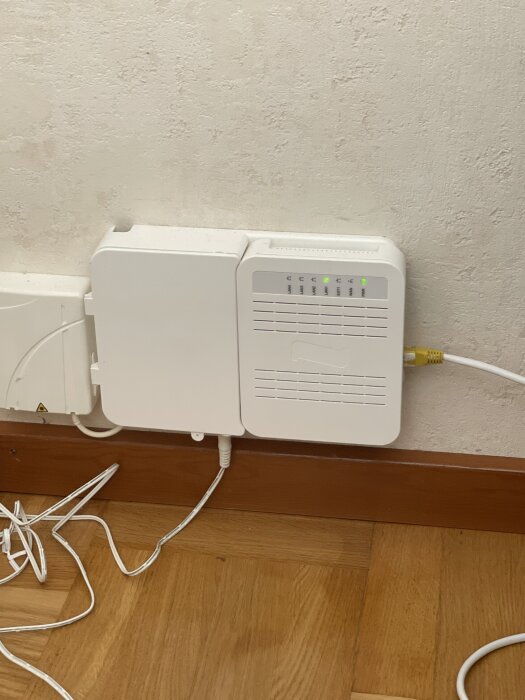 Vit internetdosa och router med blinkande lampor, placerad på en golvlist med kablar anslutna.