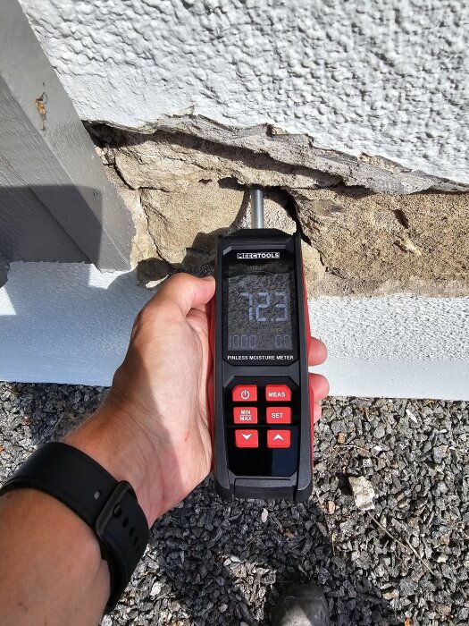 Användare mäter fukt i husfasad med fuktmätare, omgiven av skadad puts och sten.