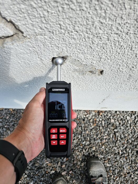 Fuktmätare används på skadad vägg för att mäta fuktnivåer, omgiven av grus och visar 12,2% på skärmen.