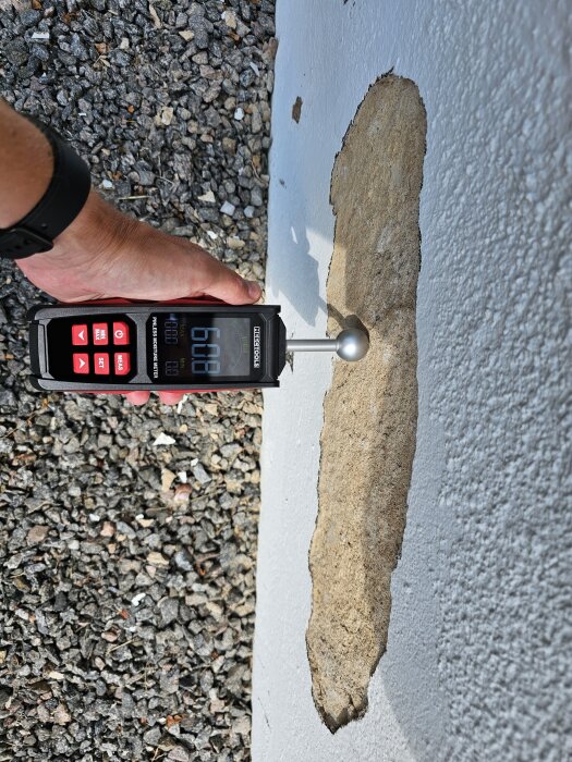 En person använder en fuktmätare på en skadad yta av en vit vägg, omgiven av grus.