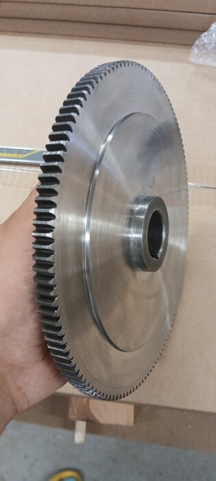 En person håller ett stort kugghjul i metall, cirka 200mm i diameter, tillverkat av förhärdat stål, del av ett byggprojekt.