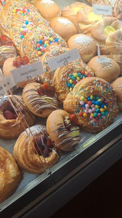 Bröd- och bakelseutbud på TM Bagarstuga, inklusive jordgubbarlyx och godisbulle med strössel och godis i ett kaféskyltfönster.