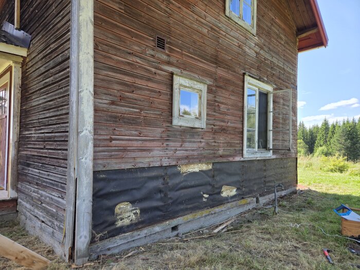 Gammal fasadvägg med avlägsnade nedre panelbrädor, exponerad vindpapp och verktyg på marken, omgiven av gräs och skog.
