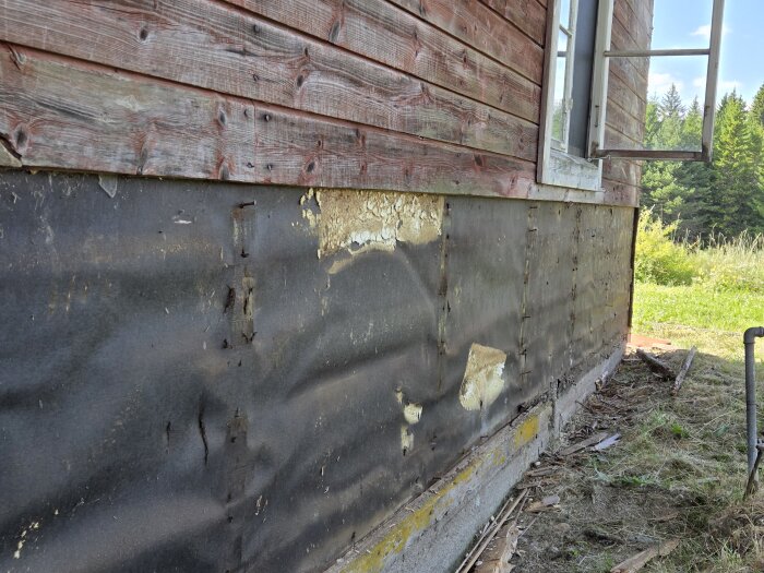 Nedersta delen av en torpvägg med avlägsnad panel, blottar skadad vindpapp och omålad yta ovan betonggrund.