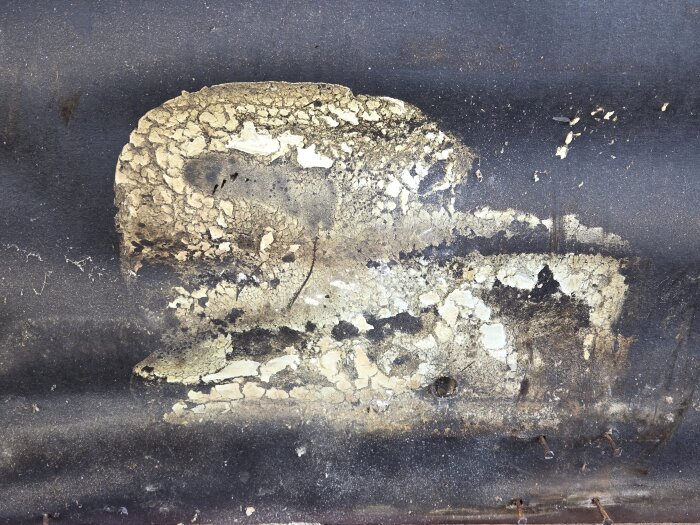 Flagnande och sprucken färg på träfasad med delvis avskalad yta. Bakgrunden är mörk, och några rostiga spikar syns längst ner.