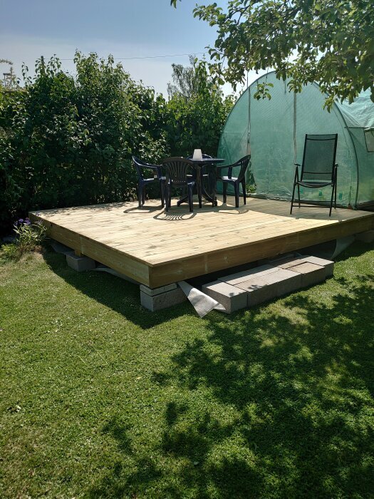 Friliggande trädäck med plaststolar och bord i en trädgård, nära ett växthus och buskar.