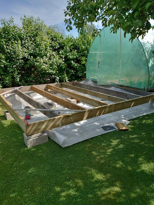 Friliggande byggprojekt med träram och betongblock på gräsmatta, grönt skyddsnät i bakgrunden.