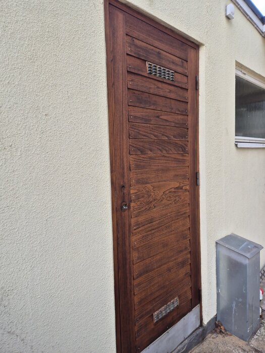 Brun träytterdörr med horisontella paneler och ventil monterad på en ljus vägg, bredvid ett silverfärgat elskåp.