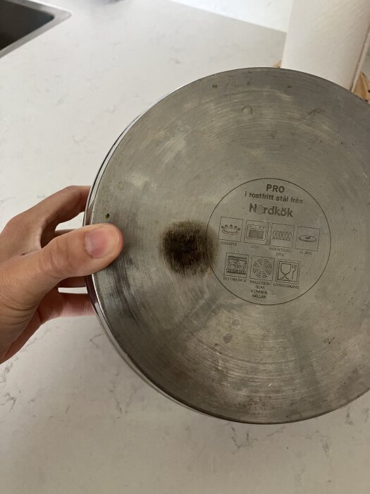 Undersidan av en kastrull med en mörk överhettad fläck i mitten; text och symboler synliga på metallen.