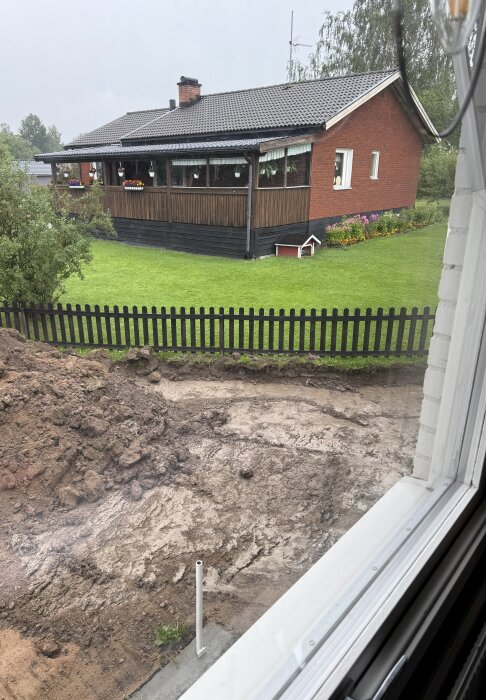Grävd uppfart utanför ett hus, med jordhögar och borttagna markplattor, omgiven av grön gräsmatta och staket.
