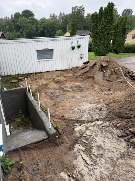Uppfart grävd ner till rätt nivå, gamla markplattor och jord borttagna. Grävt område vid vit byggnad, redo för nästa byggfas.