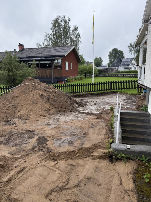 Grävd uppfart med borttagna markplattor, gräsmatta borta, redo för byggfas. Staket och hus i bakgrunden, svenska flaggan synlig.