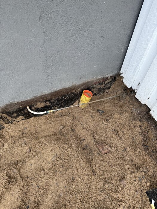 Grävd uppfart med synlig markyta och en gul kabelskyddsrör vid en grå husvägg och vitt plank.