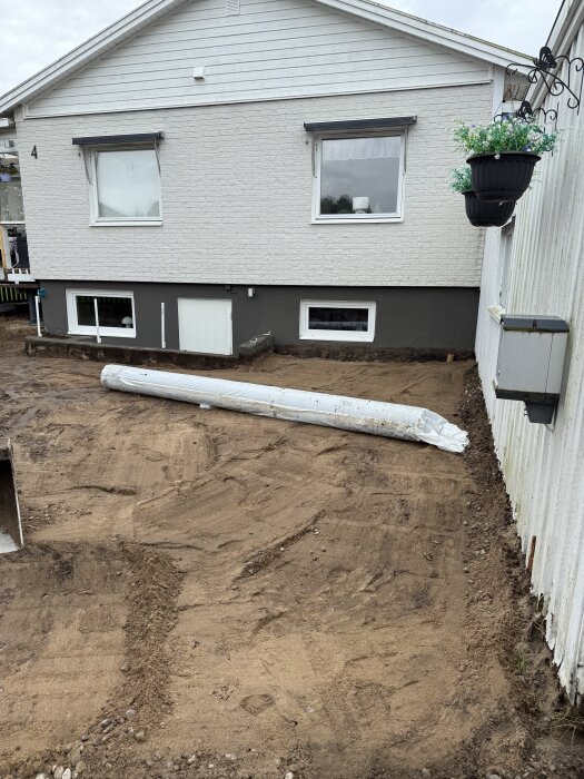 Uppfarten vid ett hus är nygrävd; marken är jämnad med synligt jordlager och en rulle med markduk ligger på marken. En vit husvägg syns i bakgrunden.